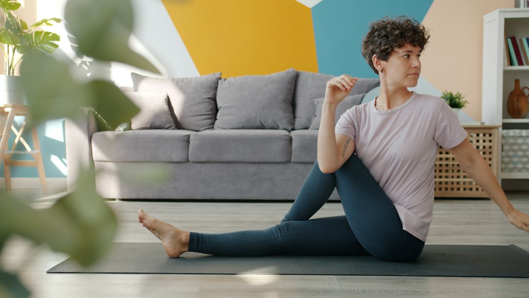 Woman with short curly hair doing a seated stretch on a mat in a bright living room