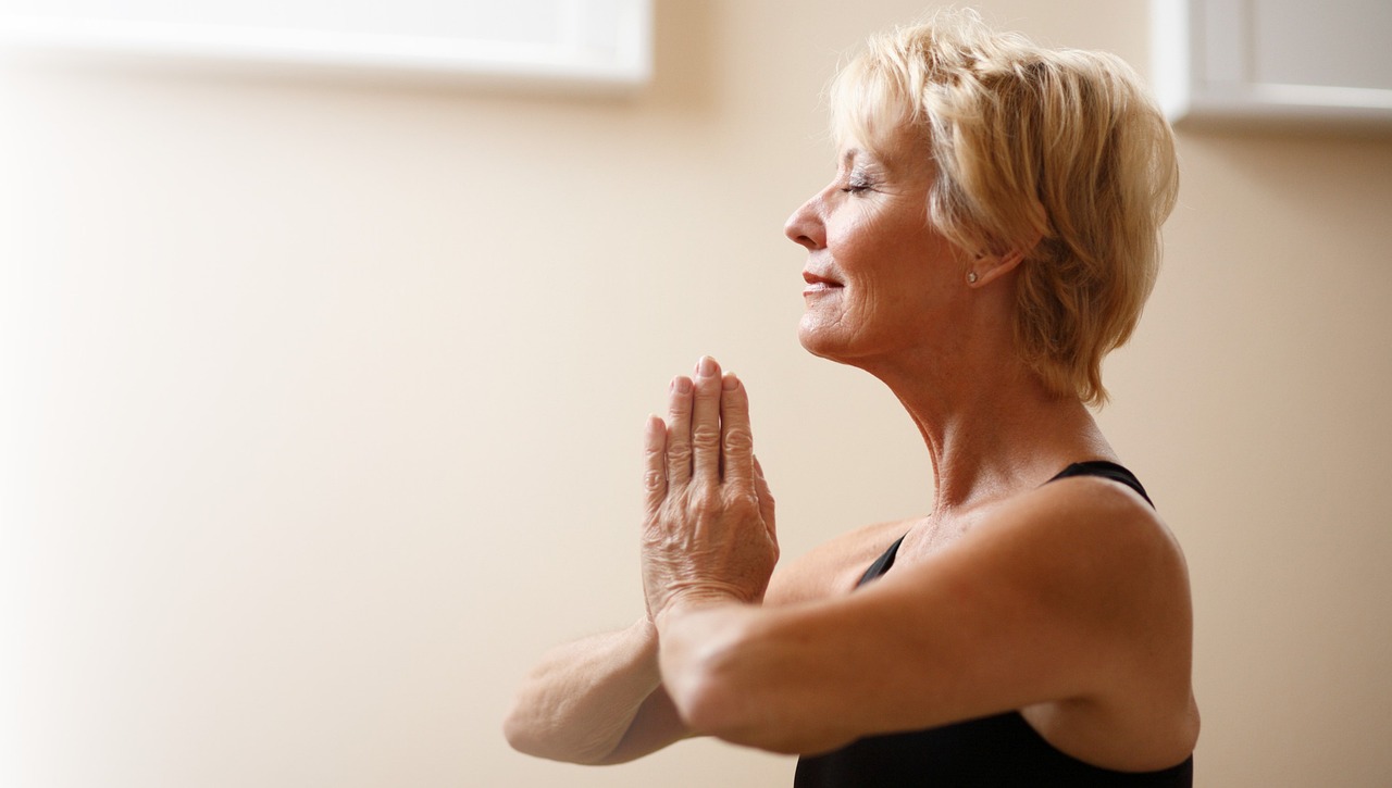A smiling older woman with eyes closed, hands pressed together, breathing calmly in a softly lit room
