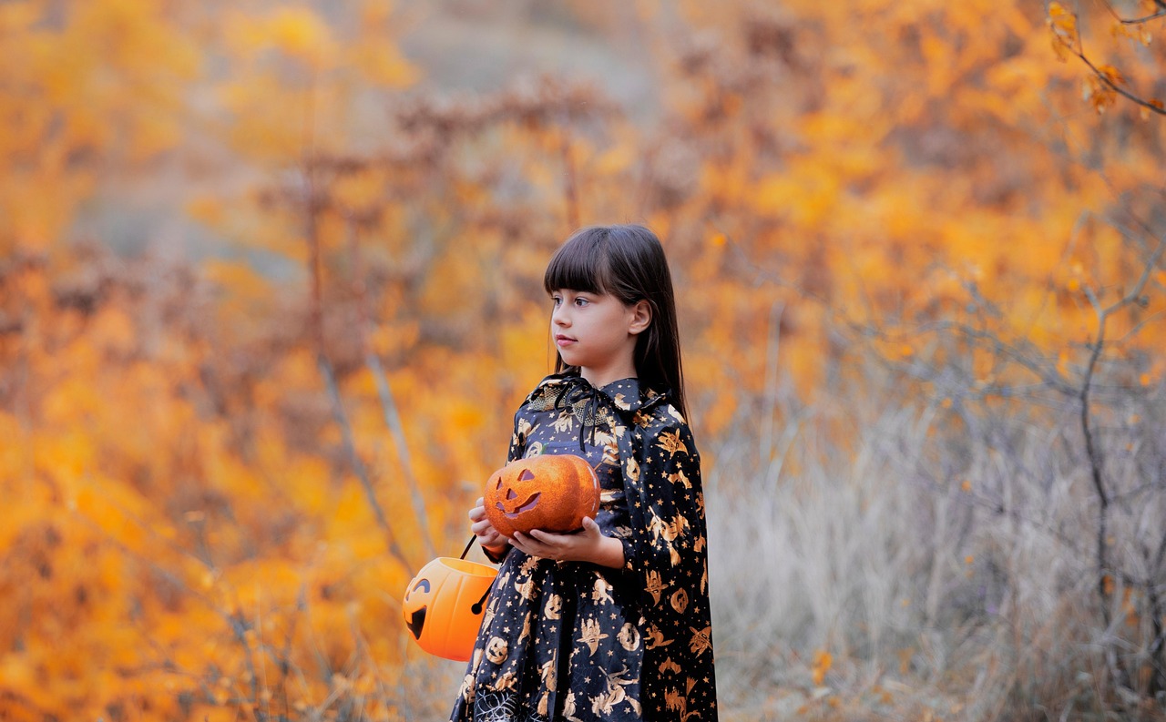 Child in a witch costume holding a jack-o'-lantern candy pail in an autumn field
