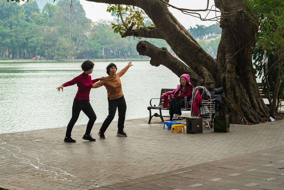 Two women practicing slow tai chi movements on a lakeside path under a large old tree while a friend watches from a bench