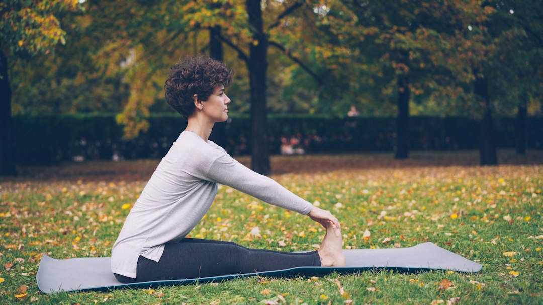 A woman seated on a mat in an autumn park, reaching toward her foot in a hamstring stretch
