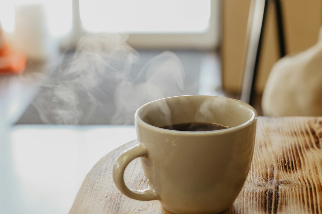 A steaming cup of tea on a wooden table beside a bright window on a cool morning