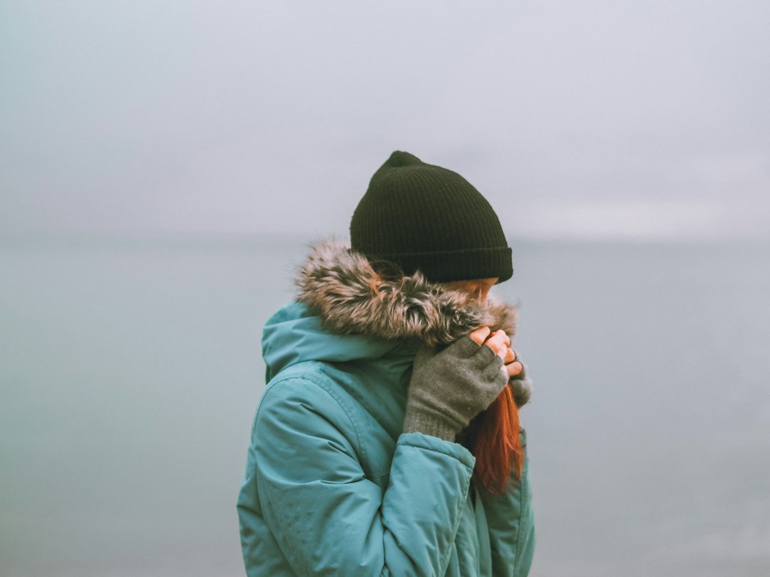 A person in a teal parka, black beanie, and gloves holding the collar closed against gray weather