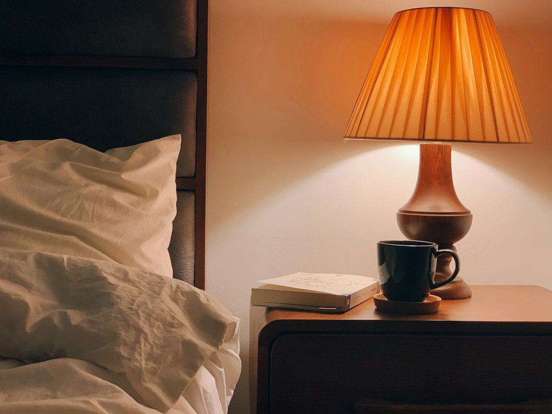 A warm bedside lamp glowing over a nightstand with a book and a dark mug next to rumpled white bedding